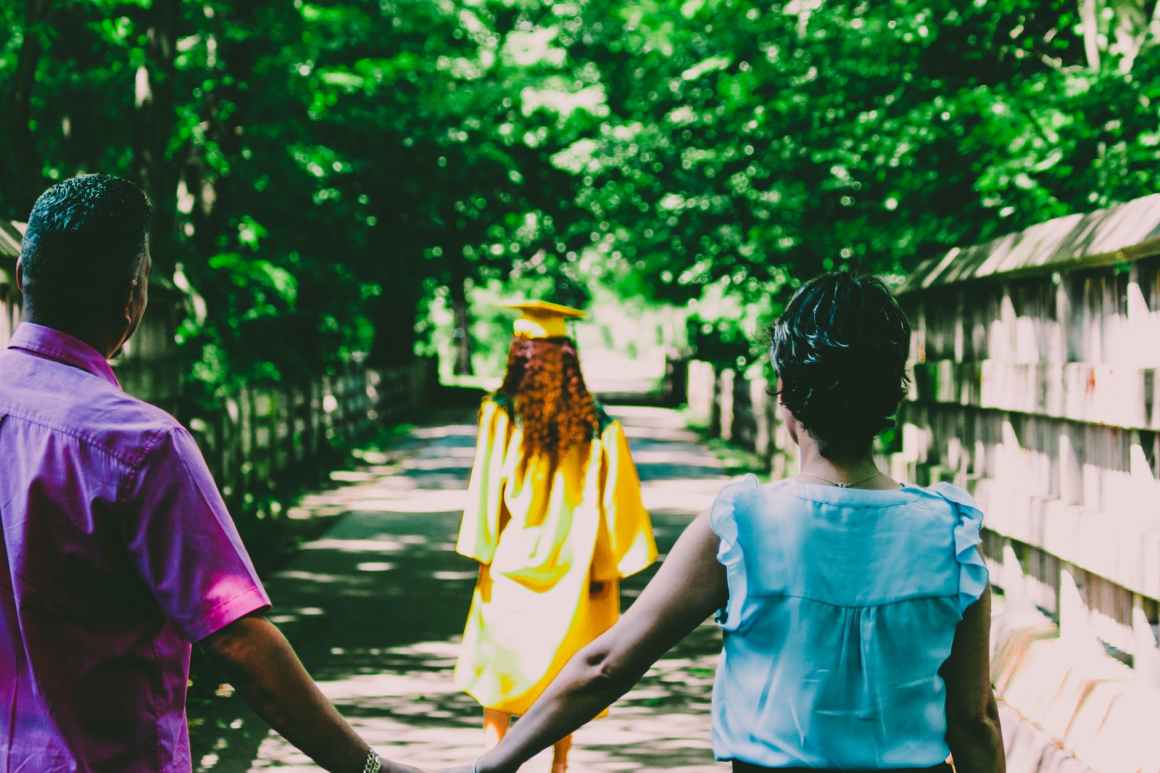 A graduate in a yellow gown walking a tree-lined path with two children